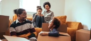 Young family of four smile and laugh together in a living room full of moving boxes.